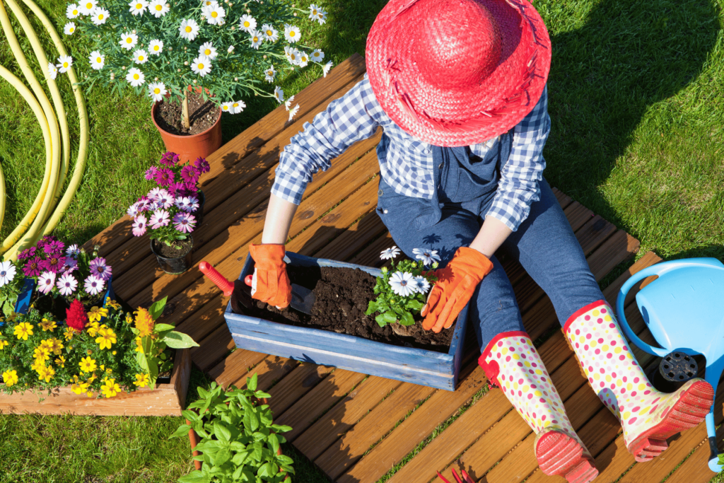 A woman with a red hat is sitting on a deck gardening and is surrounded by other flower pots